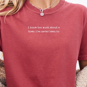 May include: A short-sleeved, crew-neck t-shirt in a muted brick red color. The center of the chest features white text that reads, I know too much about a town I've never been to. The shirt is paired with light blue denim shorts and a beige cardigan.