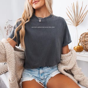 May include: A charcoal gray short-sleeve t-shirt featuring the white text "one ball python was never the plan" printed on the upper chest. The shirt has a crew neckline and a relaxed fit, paired with light blue denim shorts and a beige cardigan.