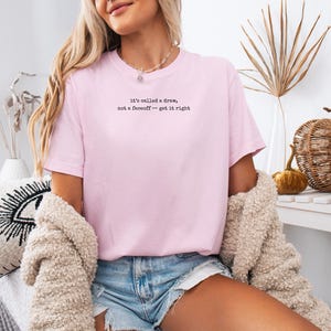 May include: A light pink short-sleeved t-shirt featuring black text printed on the chest that reads, "it's called a draw, not a faceoff -- get it right." The shirt has a crew neckline and a standard fit, paired with light blue denim shorts.