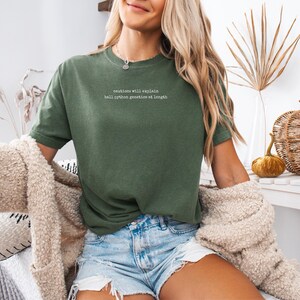 May include: An olive green short-sleeve t-shirt featuring white text on the chest that reads: "cautious will explain ball python genetics at length". The shirt has a crew neckline and is paired with light blue denim shorts and a textured beige cardigan.