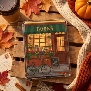Może przedstawiać: Przezroczyste etui na tablet z ilustracją księgarni, markiza z napisem "BOOKS". Zielony rower stoi przed wejściem. Jesienne liście i dynia.
