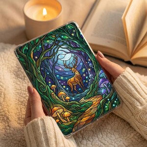 Può includere: Una custodia trasparente per telefono con una colorata scena di foresta con un cervo, funghi e una luna piena. L'opera d'arte è in stile vetrata, con verdi, blu e arancioni vibranti. Una candela accesa e un libro aperto sono sullo sfondo.