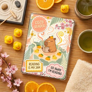 May include: A clear tablet case featuring a cartoon capybara reading a book in a bath with rubber ducks and oranges. Text includes "Cozy Reader", "Reading is my Jam", and "Go Away I'm Reading". The case is surrounded by oranges, tea, and zen garden.