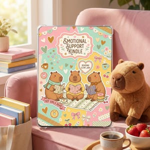 May include: A clear Kindle case with a colorful design featuring three cartoon capybaras reading books. The case has the text "EMOTIONAL SUPPORT KINDLE", "READING IS MY JAM", and "HOT GIRLS READ". The background includes hearts, bows, and donuts.