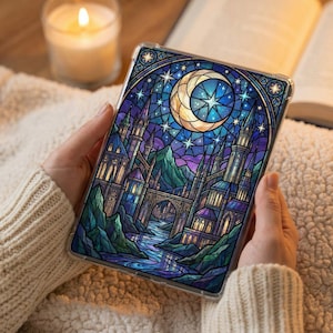 May include: A tablet case featuring a stained-glass design with a crescent moon, stars, and a castle scene in shades of blue, purple, and gold. The case is held against a cozy, cream-colored blanket, with a lit candle and an open book in the background.