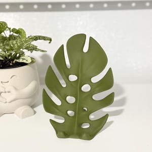 Puede incluir: Un soporte para teléfono de color verde oliva mate con forma de hoja de monstera, que presenta recortes y perforaciones características. El soporte cuenta con una base curva diseñada para sostener un dispositivo móvil en posición vertical.