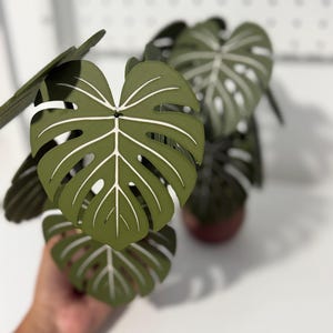 May include: A decorative artificial plant featuring multiple dark olive green monstera leaves with prominent white veins. The leaves are constructed with a layered, textured surface and are arranged to mimic the natural growth pattern of a monstera plant.