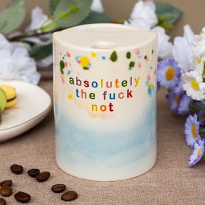 May include: A cylindrical ceramic mug with a cream-colored top and a light blue base. The surface features colorful paint splatters and the text "absolutely the fuck not" printed in multi-colored letters. The mug has a flat top with a small hole.