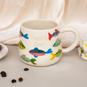 Puede incluir: Una taza de cerámica de color crema con un patrón pintado a mano de peces coloridos en tonos azul, verde, rojo y amarillo. El texto "Frank's Dad" está grabado en el centro de la taza. Tiene un cuerpo redondeado y un asa lateral resistente.