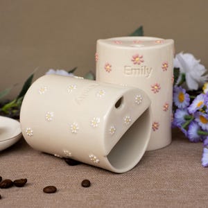 Könnte beinhalten: Zwei zylindrische Keramikbecher in Cremefarben, jeweils mit einem Muster aus kleinen erhabenen Gänseblümchen und dem eingeprägten Namen Emily an der Seite. Die Becher haben eine schräge Öffnung mit einem kleinen runden Loch.