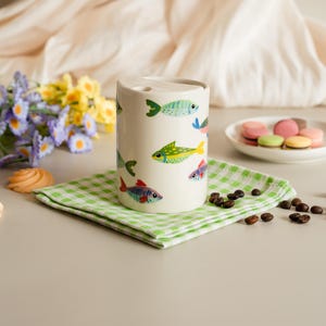 May include: A cylindrical ceramic toothbrush holder with a white background, decorated with colorful fish illustrations. The holder sits on a green and white checkered napkin, with coffee beans and macarons nearby.