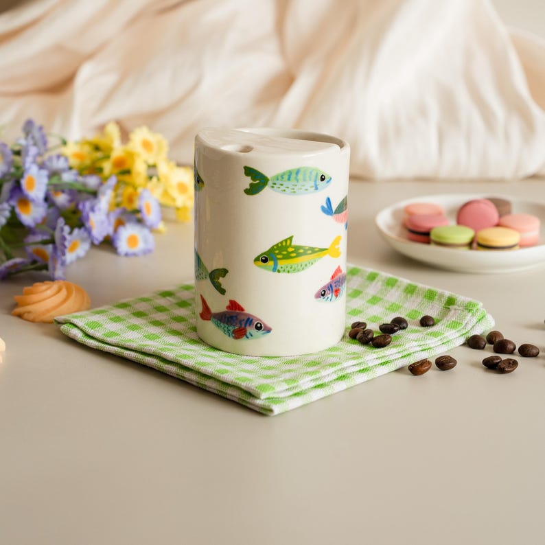 May include: A cylindrical ceramic toothbrush holder with a white background, decorated with colorful fish illustrations. The holder sits on a green and white checkered napkin, with coffee beans and macarons nearby.