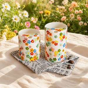 Puede incluir: Dos recipientes cilíndricos de cerámica blanca con coloridos estampados florales. Los recipientes están decorados con flores rojas, amarillas, azules y verdes. Se encuentran sobre un paño estampado en blanco y negro con granos de café. El fondo es un jardín borroso.