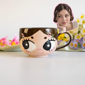 May include: A ceramic mug featuring a stylized face with brown hair, large black eyes with white star highlights, and pink cheeks. The mug includes a dark brown handle and decorative hair clips painted on the hair section, mirroring the person shown.