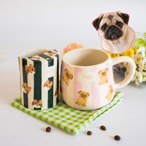 Puede incluir: Una taza cilíndrica con rayas verde oscuro y blancas con caras de gato y el nombre Jonny, junto a una taza redondeada con rayas rosa claro y blancas con ilustraciones de carlinos y el nombre Kelly.