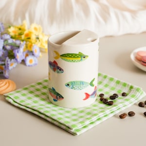 May include: A cream-colored ceramic utensil holder with a curved top and a slot. The holder is decorated with colorful fish illustrations. It sits on a green and white checkered napkin, with coffee beans scattered around.