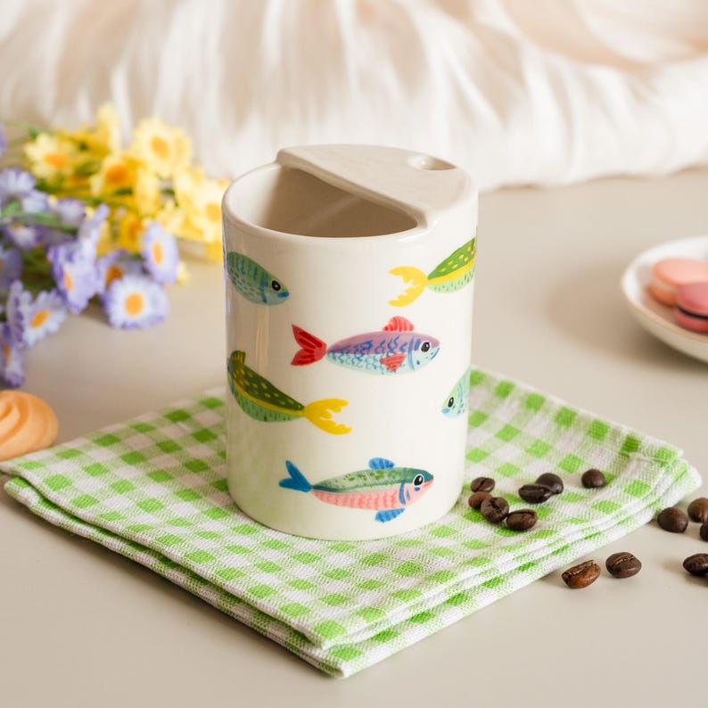 May include: A white ceramic cup with a handle, decorated with colorful fish illustrations. The cup sits on a green and white checkered napkin. Coffee beans are scattered around the napkin.