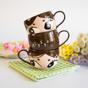 May include: A stack of three brown ceramic mugs with handles. The top and bottom mugs feature a hand-painted face with large eyes and star accents. The middle mug is solid brown with the name Alyssa written in white cursive script lettering.