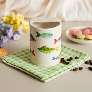 May include: A cylindrical ceramic container with a handle, decorated with colorful fish illustrations. The container sits on a green and white checkered napkin. Coffee beans and a plate of macarons are also present.