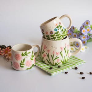 May include: Three cream-coloured ceramic mugs with hand-painted floral designs and personalised names. One mug features pink roses and the name Amber, another shows colourful wildflowers and the name Carly, and the third displays pink tulips and the name Chloe.