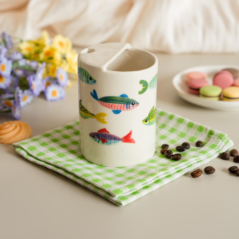 May include: A white ceramic mug decorated with colorful fish illustrations. The mug has a unique handle design and sits on a green and white checkered napkin. Coffee beans and macarons are also visible.