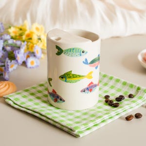 May include: A white ceramic tumbler with a curved top and a series of colorful fish illustrations. The fish are in shades of blue, green, yellow, and red. The tumbler sits on a green and white checkered napkin.