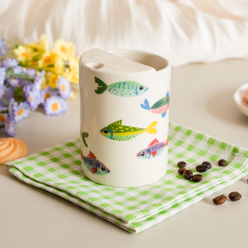 May include: A white ceramic tumbler with a curved top and a series of colorful fish illustrations. The fish are in shades of blue, green, yellow, and red. The tumbler sits on a green and white checkered napkin.