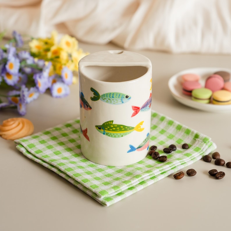May include: A cylindrical ceramic utensil holder with a handle, decorated with colorful fish illustrations. The holder sits on a green and white checkered napkin, with coffee beans and a plate of macarons in the background.