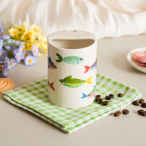 May include: A cylindrical ceramic container with a rectangular opening at the top, decorated with colorful fish illustrations. The container sits on a green and white checkered napkin. Coffee beans and flowers are in the background.