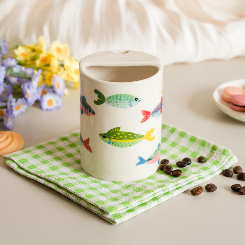 May include: A cylindrical ceramic container with a rectangular opening at the top, decorated with colorful fish illustrations. The container sits on a green and white checkered napkin. Coffee beans and flowers are in the background.