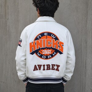 May include: A white leather varsity jacket featuring a large circular New York Knicks logo on the back with orange and navy blue lettering. The word KITH is centered in the logo. The word AVIREX is embroidered below in navy blue. A small American flag patch is on the right sleeve.