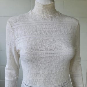 May include: A cream-colored long-sleeve top featuring a high mock neck collar and an intricate, repeating diamond-shaped knit pattern throughout the fabric. The garment is constructed with a textured, open-weave design that creates a consistent geometric aesthetic across the chest, shoulders, and sleeves.
