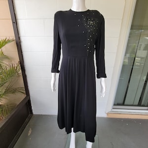 May include: A long black dress with long sleeves and a round neckline. The dress features a decorative embellishment on the upper left chest area with small white beads. The dress is displayed on a white mannequin.