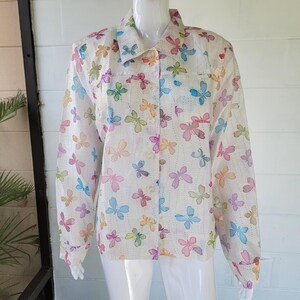 May include: A long-sleeved button-down shirt in white, featuring a repeating pattern of butterflies in shades of blue, pink, yellow, and green, along with vertical dotted lines. The shirt has a classic collar and two chest pockets on the front.