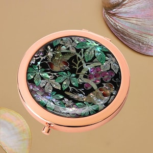 May include: A circular, rose gold compact mirror featuring an inlaid design of birds, grapes, and leaves. The design is made of iridescent mother-of-pearl against a black background. The mirror has a small clasp for opening and closing.