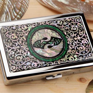 May include: A silver cigarette case with a black, mother-of-pearl inlaid design. The design features a central green circle with two birds and a decorative border, surrounded by swirling patterns.
