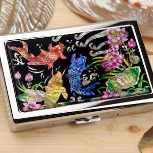 May include: A silver business card holder with a black inlay depicting colourful koi carp and floral designs. The carp are red, blue, and yellow, with pink and green flowers. The holder has a secure clasp.