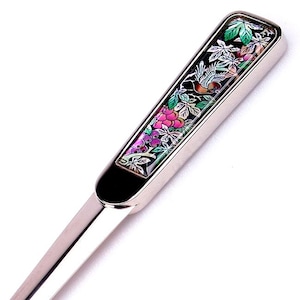 May include: A silver letter opener with a decorative handle. The handle features a rectangular inlay with a floral and bird design in shades of pink, green, and black. The blade is long and slender.