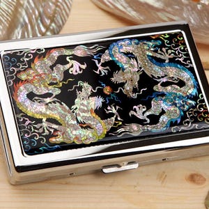 May include: A rectangular, silver-toned case with a black top featuring a dragon design. The dragons are depicted in iridescent colors, including silver, blue, and gold, against a black background. The case has a shiny, metallic finish.