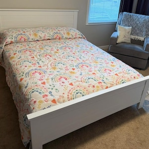 Può includere: Letto in legno bianco con una trapunta colorata con arcobaleni, cuori e motivi floreali. Il design della trapunta include sfumature di rosa, giallo, blu e arancione. Sullo sfondo una poltrona grigia con una coperta e un cuscino.