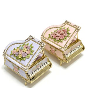 Puede incluir: Dos pianos de cola en miniatura decorativos en colores pastel. Uno es lavanda, el otro rosa, ambos con detalles dorados. Cada uno presenta un arreglo floral en la tapa y un teclado. Probablemente sean cajas de música o artículos decorativos.