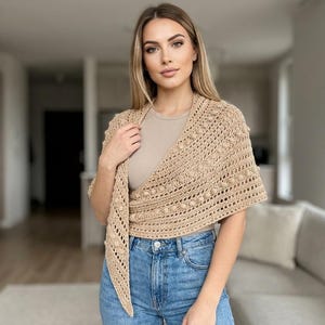 May include: A beige, crocheted shawl with a textured, bobble stitch design. The shawl is draped over the shoulders and tied in the front. The garment is worn over a neutral-colored top and paired with blue jeans.