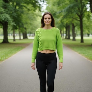 May include: A woman wearing a lime green cropped sweater with a braided cable design on the left shoulder and sleeve. She is wearing black leggings and walking on a paved path lined with trees.
