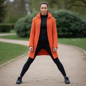 Puede incluir: Un cárdigan largo naranja con cuello ancho, usado sobre una camiseta negra y leggings negros. La persona lleva zapatos negros y está de pie en un camino en un parque. El cárdigan es una prenda llamativa.