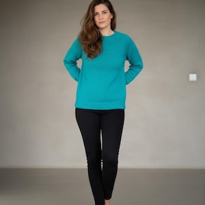 Può includere: Una donna indossa un maglione turchese e pantaloni neri. Il maglione ha un girocollo e maniche lunghe. I pantaloni sono aderenti. La donna ha lunghi capelli castani e si trova in una stanza con pareti neutre.