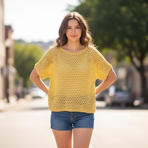May include: A woman wearing a yellow, open-weave crochet top with short sleeves and blue denim shorts. The top has a round neckline. The background shows a city street with buildings and trees.