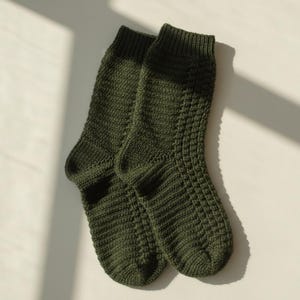 May include: A pair of olive green knitted socks. The socks are made of a textured knit pattern and are lying on a light-colored surface. The socks are in a natural light setting.