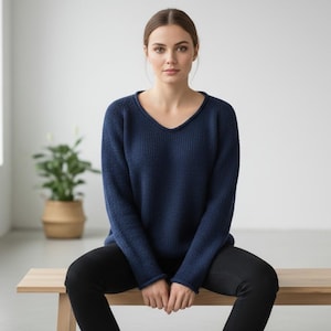 May include: A navy blue sweater with a V-neck. The long-sleeved knitwear is worn with black pants. The person is sitting on a light-colored wooden bench. A potted plant is in the background.