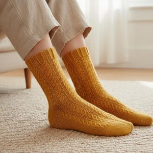 May include: A pair of mustard yellow knitted socks with a cable knit pattern. The socks are worn by a person sitting on a beige carpet. The socks have a ribbed cuff and a textured design.