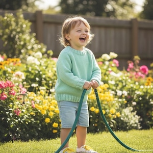 May include: A young child wearing a light blue sweater and denim shorts laughs while holding a green garden hose. The child is standing on green grass in front of a colorful flower garden.